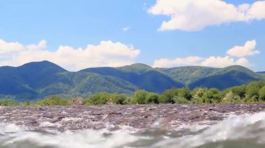 Serin yaz manzarası parçalı bulutlu bir gökyüzünün altında yemyeşil bir dağ sırasına akan kayalık bir dağ nehrine ev sahipliği yapar..
