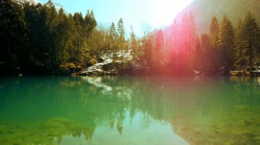 Güneşli çam ağaçları ve karla kaplı dağ yamaçlarını yansıtan kristal berraklığında turkuaz suyu olan sakin alp gölü..