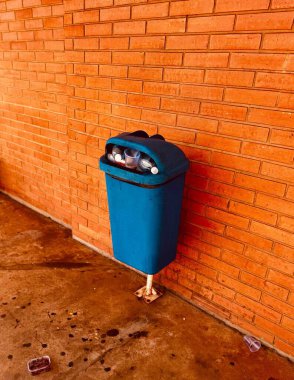 Blue trash can full of trash on the floor leaning against an exposed brick wall  clipart