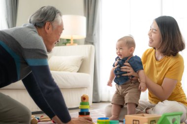 Küçük bir çocuk, anne ve büyükbabasıyla oturma odasında oyuncaklarla neşeyle oynuyor.