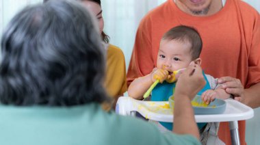 Küçük bir çocuk ilk yemeğini yiyor, ebeveynler ve büyük anne ve babalar coşkuyla alkışlıyorlar.