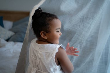 Küçük bir kız yatak odasında perdelerle saklambaç oynuyor.