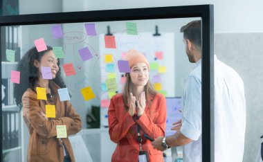 Çok ırklı genç iş adamları ofisteki cam duvara yazı yazarak bir proje üzerinde beyin fırtınası yapıyorlar..