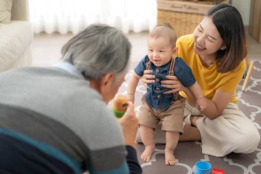 Küçük bir çocuk, anne ve büyükbabasıyla oturma odasında oyuncaklarla neşeyle oynuyor.