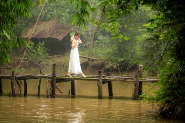 Beyaz elbiseli genç bir kadın nehrin karşısındaki eski ahşap köprüde yürüyor. Sabah sisli bir hava var.