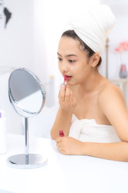 Kafasına ve vücuduna havlu sarılmış, duştan sonra makyaj yapan Asyalı genç bir kadın.