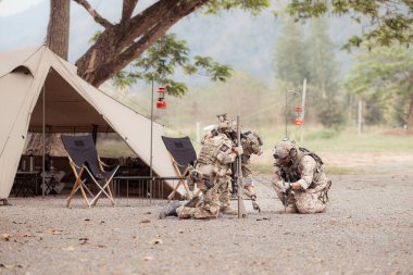 Kamuflaj üniformalı bir grup asker kamp çadırının önünde silah tutuyor, savaş eğitimi için plan yapıyor ve hazırlanıyor..