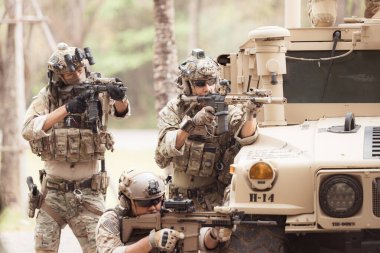 Kamuflaj üniformalı askerler, askeri haberleşme aracının yan tarafında saklanarak silahlarını ateşe hazır tutuyorlar.