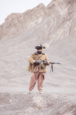 Bir Taliban askeri elinde makineli tüfekle çöldeki dağ manzarasında duruyor.