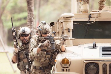Kamuflaj üniformalı askerler, askeri haberleşme aracının yan tarafında saklanarak silahlarını ateşe hazır tutuyorlar.