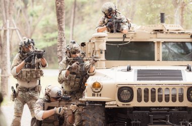 Kamuflaj üniformalı askerler, askeri haberleşme aracının yan tarafında saklanarak silahlarını ateşe hazır tutuyorlar.