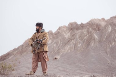 Bir Taliban askeri elinde makineli tüfekle çöldeki dağ manzarasında duruyor.