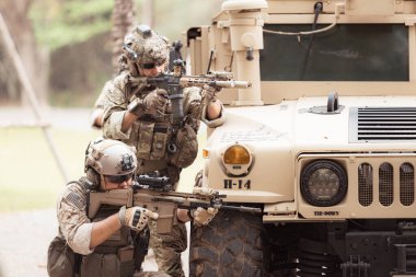 Kamuflaj üniformalı askerler, askeri haberleşme aracının yan tarafında saklanarak silahlarını ateşe hazır tutuyorlar.