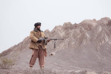 Bir Taliban askeri elinde makineli tüfekle çöldeki dağ manzarasında duruyor.