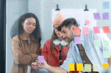 Çok ırklı genç iş adamları ofisteki cam duvara yazı yazarak bir proje üzerinde beyin fırtınası yapıyorlar..