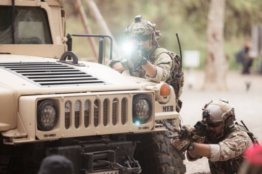 Kamuflaj üniformalı askerler, askeri haberleşme aracının yan tarafında saklanarak silahlarını ateşe hazır tutuyorlar.