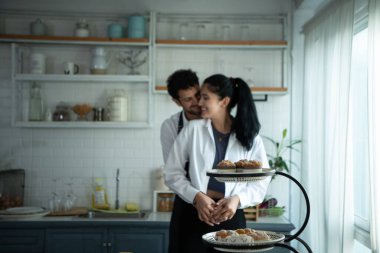 Bir koca karısıyla yemek pişirirken önlük giyer. Yemek pişirmek için mutfağa gitmeden önce karısına mutlu bir şekilde sarıldı..