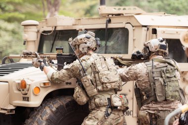 Kamuflaj üniformalı askerler, askeri haberleşme aracının yan tarafında saklanarak silahlarını ateşe hazır tutuyorlar.