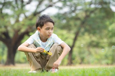Çocuk parkta oturmuş, hava kabarcığı üflüyor, çevresi yeşillik ve doğa ile çevrili.