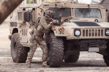 Kamuflaj üniformalı askerler, askeri haberleşme aracının yan tarafında saklanarak silahlarını ateşe hazır tutuyorlar.