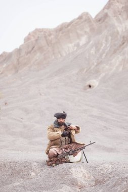 Bir Taliban askeri elinde makineli tüfekle, çöldeki dağ manzarasında.