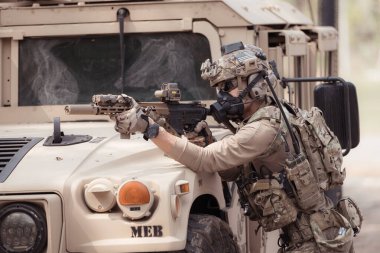 Kamuflaj üniformalı askerler, askeri haberleşme aracının yan tarafında saklanarak silahlarını ateşe hazır tutuyorlar.