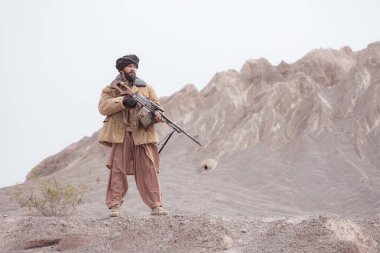 Bir Taliban askeri elinde makineli tüfekle çöldeki dağ manzarasında duruyor.