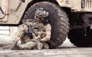 Kamuflaj üniformalı askerler, askeri haberleşme aracının yan tarafında saklanarak silahlarını ateşe hazır tutuyorlar.