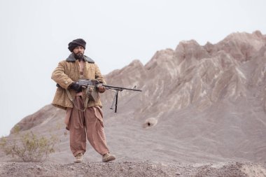 Bir Taliban askeri elinde makineli tüfekle çöldeki dağ manzarasında duruyor.