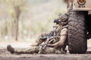 Kamuflaj üniformalı askerler, askeri haberleşme aracının yan tarafında saklanarak silahlarını ateşe hazır tutuyorlar.