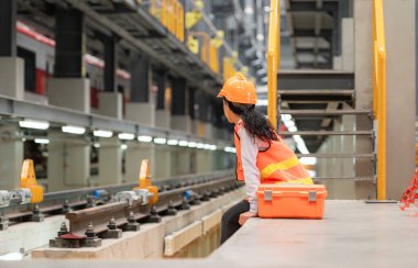Alet çantalı kadın teknisyen portresi, gökyüzü tamir istasyonundaki elektrikli tren raylarının önünde çimento kaidesinin üzerinde oturuyor..