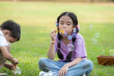 Parkta oturmuş hava kabarcığı üfleyen çocuklar çevresi yeşillik ve doğa ile çevrili.