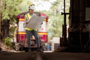 Demiryolu teknisyeninin portresi. Güvenlik yeleği ve miğferi var. Tren tamir istasyonunun planlarıyla çalışıyor.