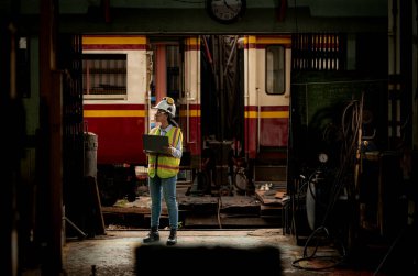 Demiryolu teknisyeninin portresi. Güvenlik yeleği ve miğferi var. Tren tamir istasyonunun planlarıyla çalışıyor.