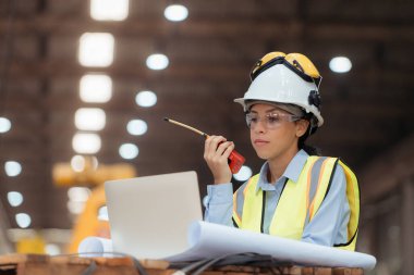 Demiryolu teknisyeninin portresi. Güvenlik yeleği ve miğferi var. Tren tamir istasyonunun planlarıyla çalışıyor.