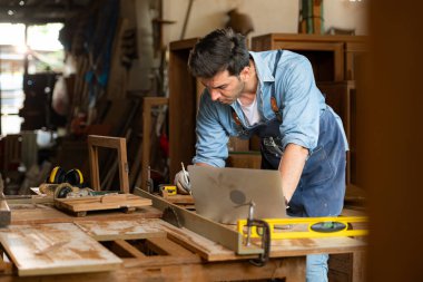 Marangoz bir marangozhanedeki atölyesinde dizüstü bilgisayarla çalışıyor.