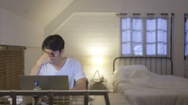 Evde fazla mesai yapan genç Asyalı adam stres ve baş ağrısını azaltmak için ilaç alıp su içiyor..