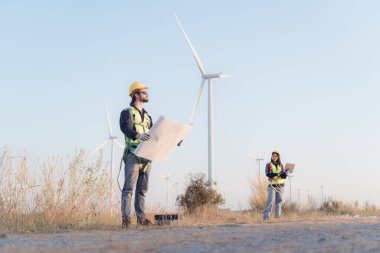 Güvenlik kaskı takan ve rüzgar türbini çiftliğinin planlarını tutan genç mühendis..