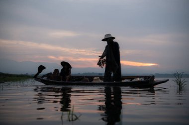 Gün doğumunda bir balıkçının silueti, bir sandalda duruyor ve yiyecek için balık yakalamak için ağ atıyor.