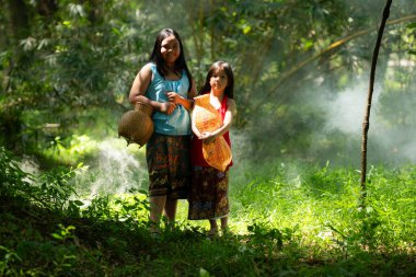 Geleneksel giyinen iki Asyalı kız yağmur ormanlarında ayakta duruyor. Büyükbabaya yakınlardaki bir gölde balık tutmasında yardım etmeden önce birlikte oynayarak eğlendiler..