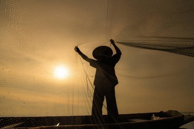 Gün doğumunda bir balıkçının silueti, bir sandalda duruyor ve yiyecek için balık yakalamak için ağ atıyor.