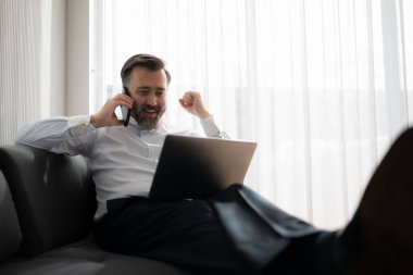 İş adamı evde kanepede otururken dizüstü bilgisayar kullanıyor ve telefonla konuşuyor.