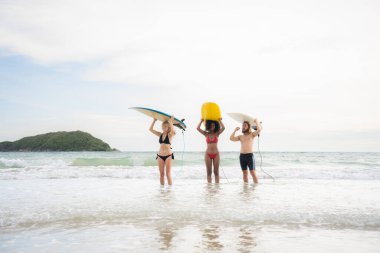 Kafalarında sörf tahtası olan iki kadın ve genç adamın arka görüntüsü ve sörf yapmak için denize girişi.