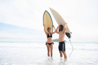 Genç bir adam ve bir kadın kafalarında sörf tahtaları tutuyorlar ve sörf yapmak için denize giriyorlar.