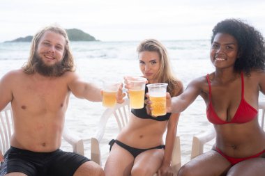 Çok ırklı bir arkadaş grubu sahilde içkili sandalyelerde oturuyor.