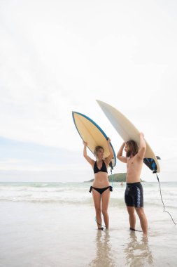 Genç bir adam ve bir kadın kafalarında sörf tahtaları tutuyorlar ve sörf yapmak için denize giriyorlar.
