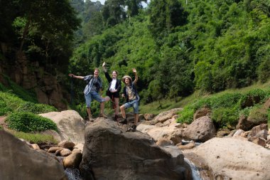 Sırt çantalı ve yürüyüş direkleri olan genç bir adam kayanın üzerinde durması için onu yukarı çekmeye yardım ediyor.
