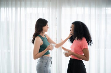 Her iki neşeli genç kadın da ellerinde kahve fincanları, ellerinde kahve fincanları ve ışığın parladığı oturma odasının beyaz perdelerinin arkasında sohbet ediyorlar.