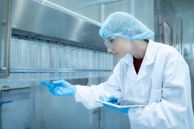 Kadın kalite kontrol işçisi içme suyu fabrikasındaki üretim hattında su şişesini inceliyor.