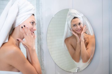 Genç ve güzel bir kadın aynanın önünde yüzüne krem sürüyor.